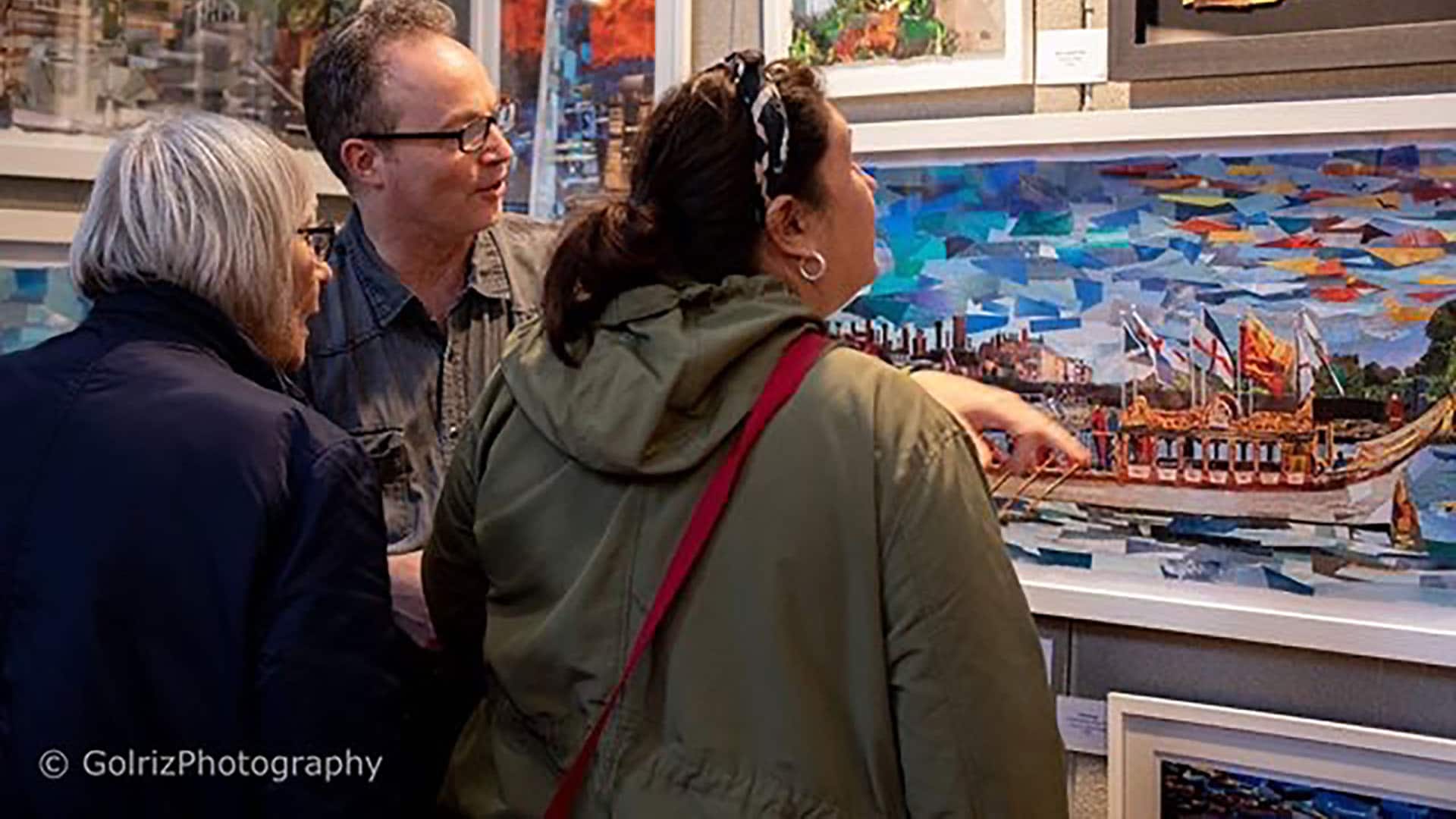 A photograph of two women and a man looking at colourful paintings, the man is pointing and explaining the painting.