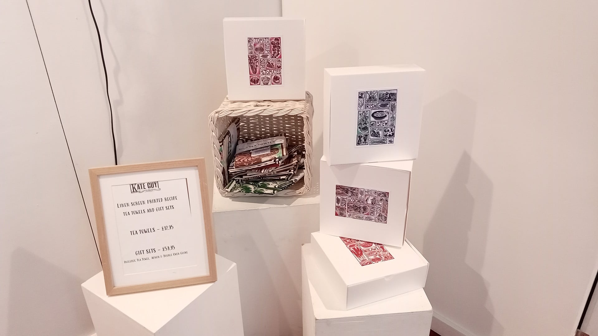 A photograph of hand crafted gifts laid out in a decorative way, with a sign stating the price of gift sets and tea towels.